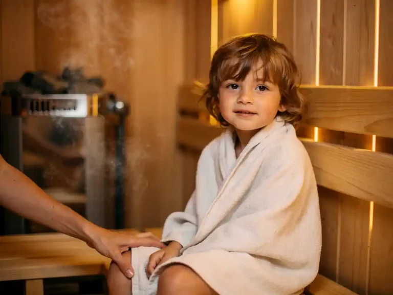 Young child wrapped in linen sitting calmly in a warm wooden home sauna, cedar bench and heater visible, parent's hand nearby.