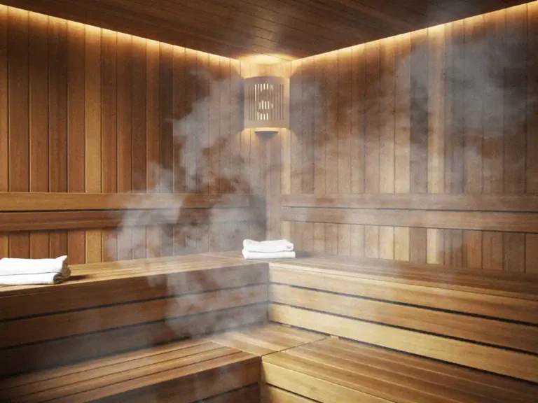 Cedar wood sauna interior with steam rising from benches, warm amber lighting, folded towels, and a mechanical vent integrated into the wall.